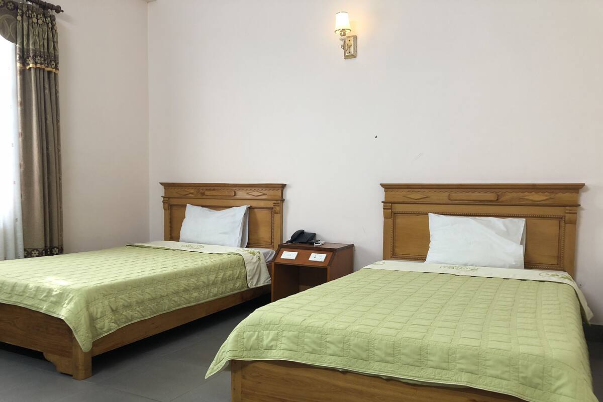 Dos camas de madera con ropa de cama verde, una mesita de noche con un teléfono y una lámpara de pared.