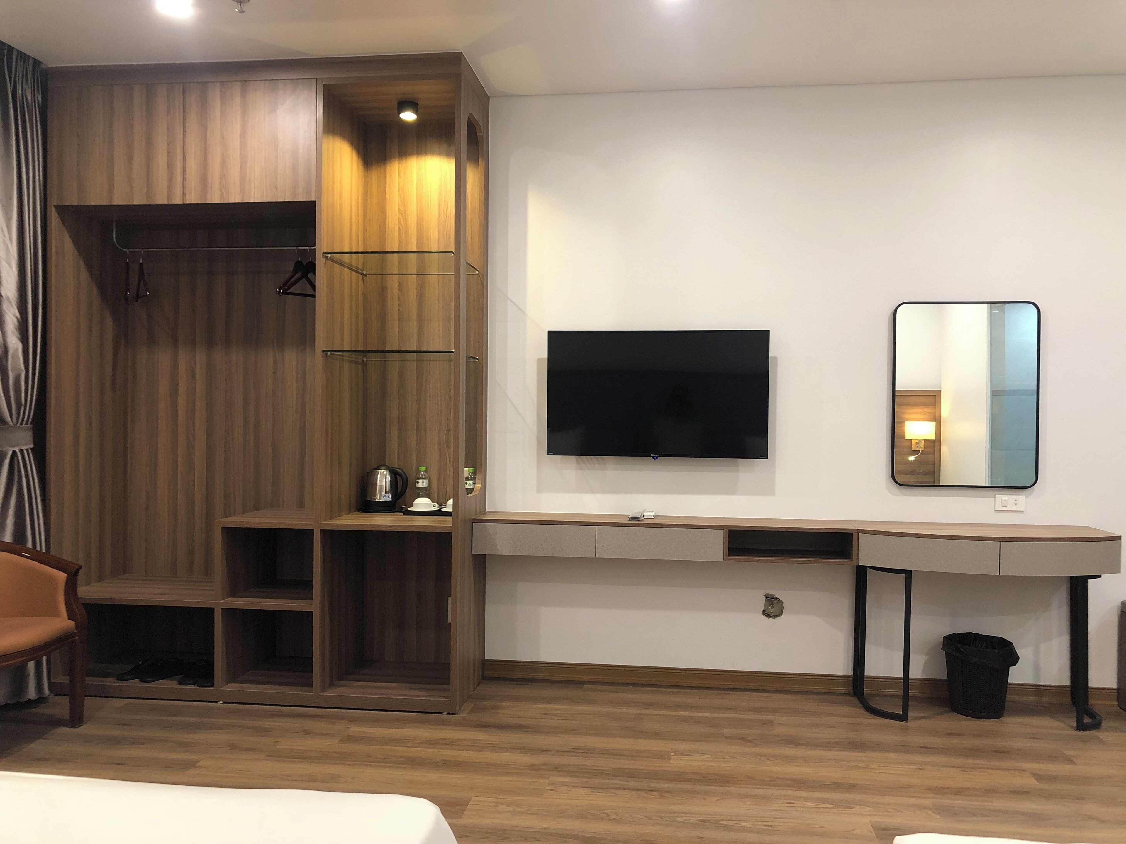 A modern hotel room with a wooden wardrobe, a flat-screen TV, a mirror, a desk, and a chair.