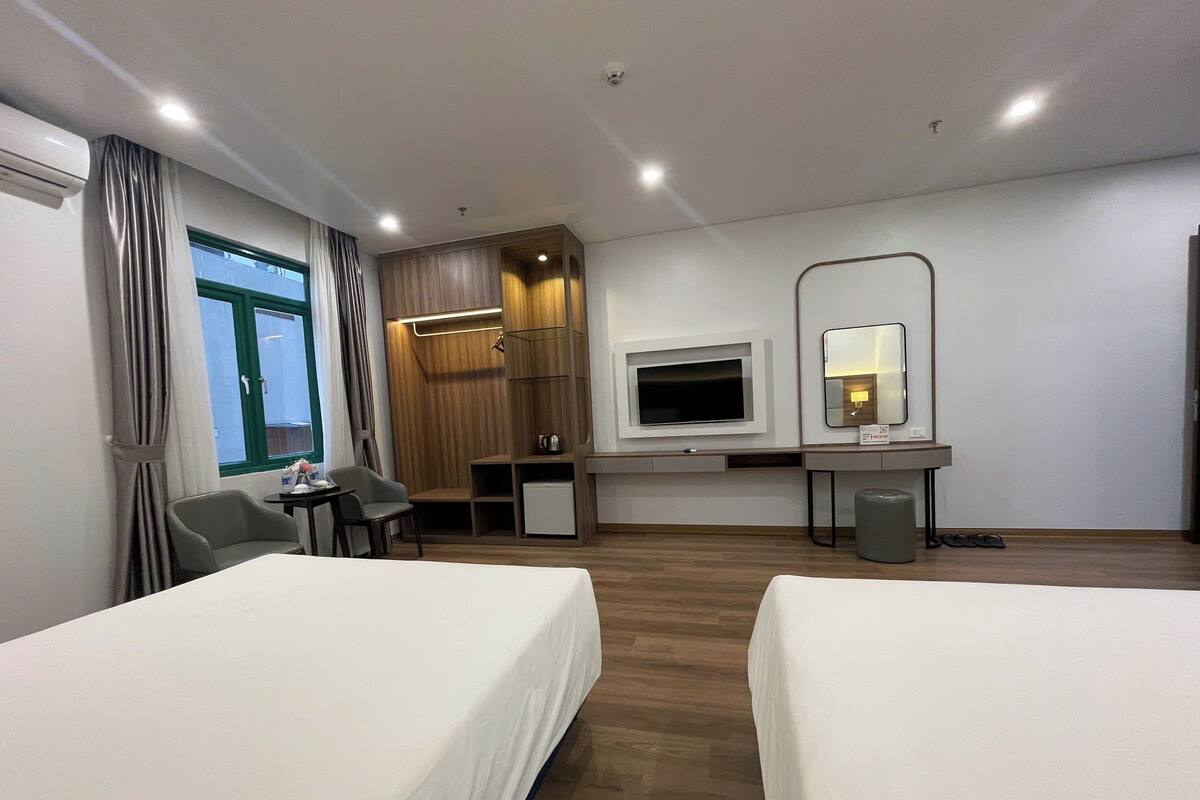Habitación de hotel con dos camas, un escritorio con televisor, una kitchenette pequeña y un ventanal grande con cortinas.