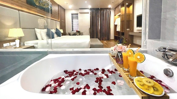 Une chambre d’hôtel moderne avec une grande baignoire remplie de pétales de roses et de morceaux de fruits, un lit, une télévision et une salle de bain équipée d’un miroir et d’un lavabo.