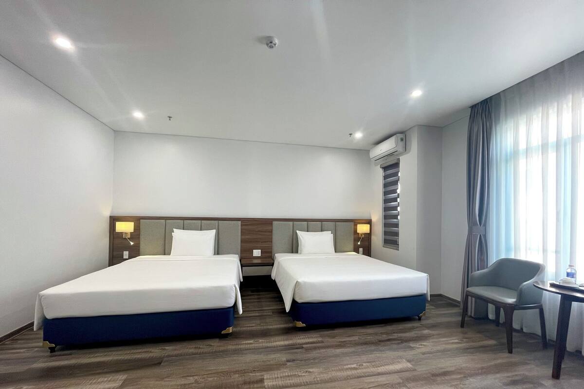Habitación de hotel con dos camas, un escritorio, una silla y un ventanal con cortinas.