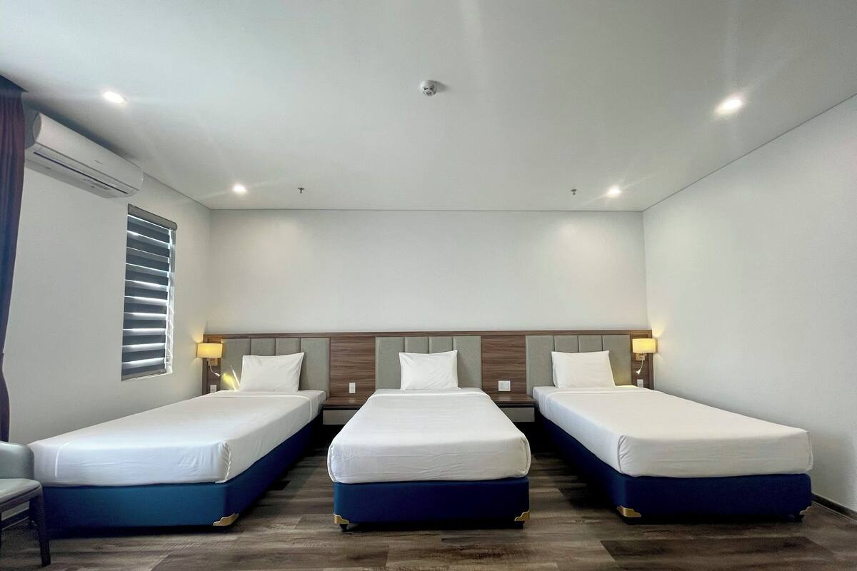 Habitación de hotel con tres camas individuales, una ventana con persianas, suelo de madera e iluminación empotrada en el techo.