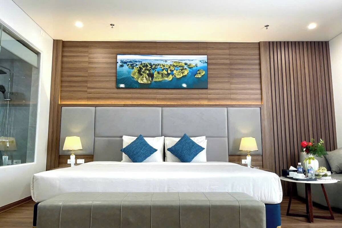 Habitación de hotel moderna con una cama grande, dos lámparas de noche, un cabecero con un cuadro y una mesita de noche con un florero, con vista a una masa de agua.