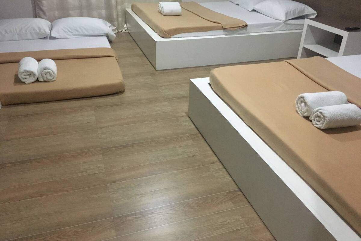 A hotel room with three beds, each with a beige mattress and white pillows. There are rolled towels on each bed. The room has a wooden floor and a white nightstand with a lamp.