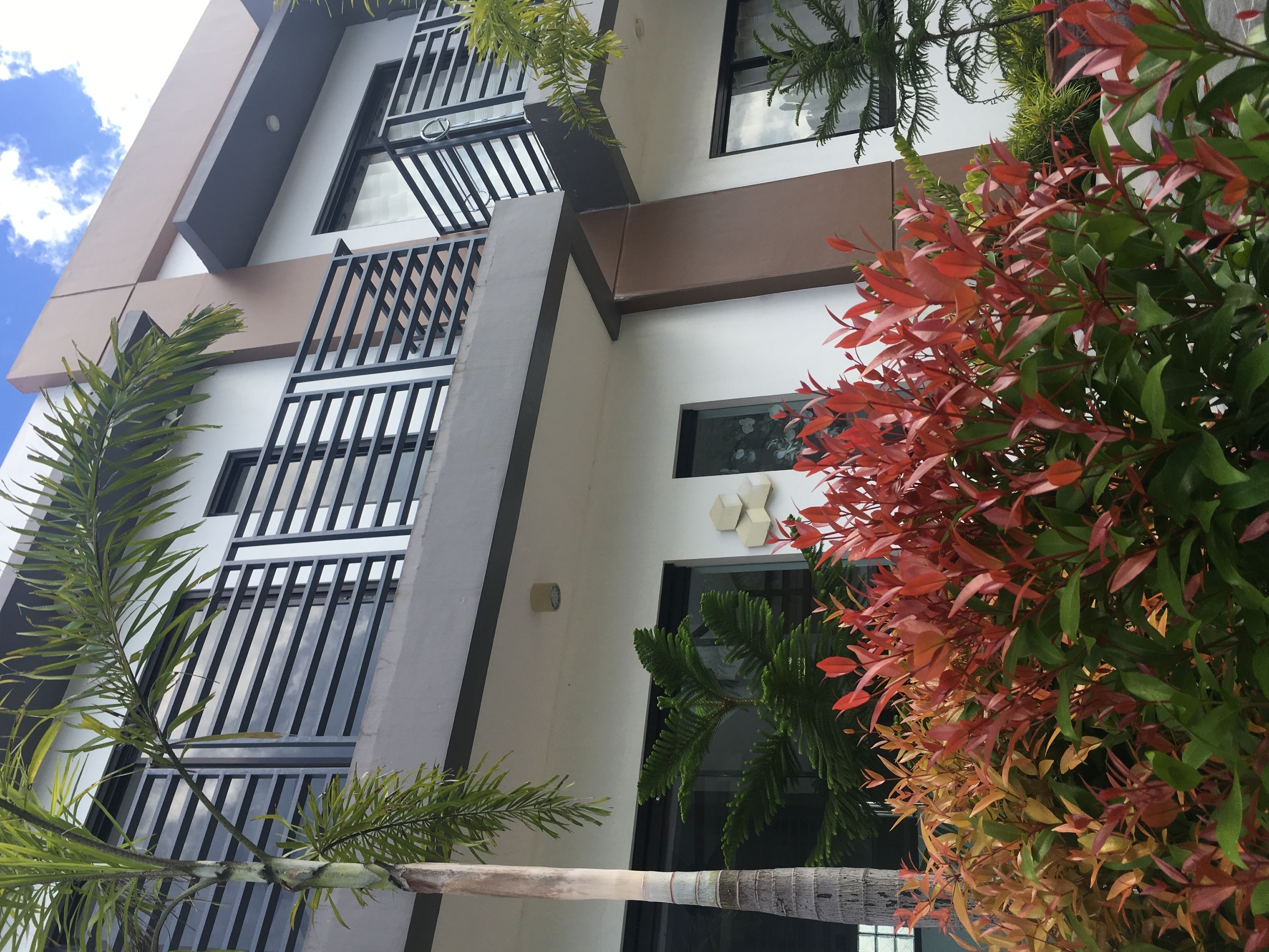 A multi-story building with balconies, large windows, and a mix of greenery including trees and shrubs.