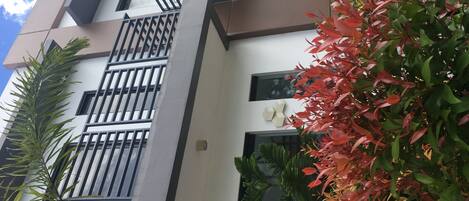 A multi-story building with balconies, large windows, and a mix of greenery including trees and shrubs.