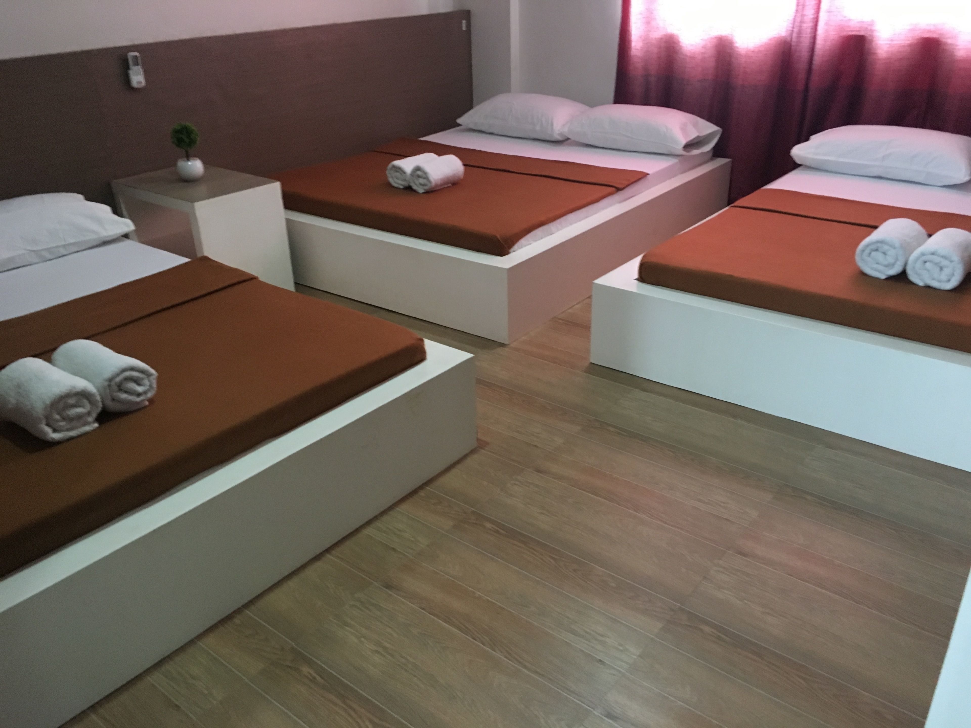 A hotel room with three beds, each with a brown mattress and white pillows. There is a small table with a plant and a white lamp on the left side.
