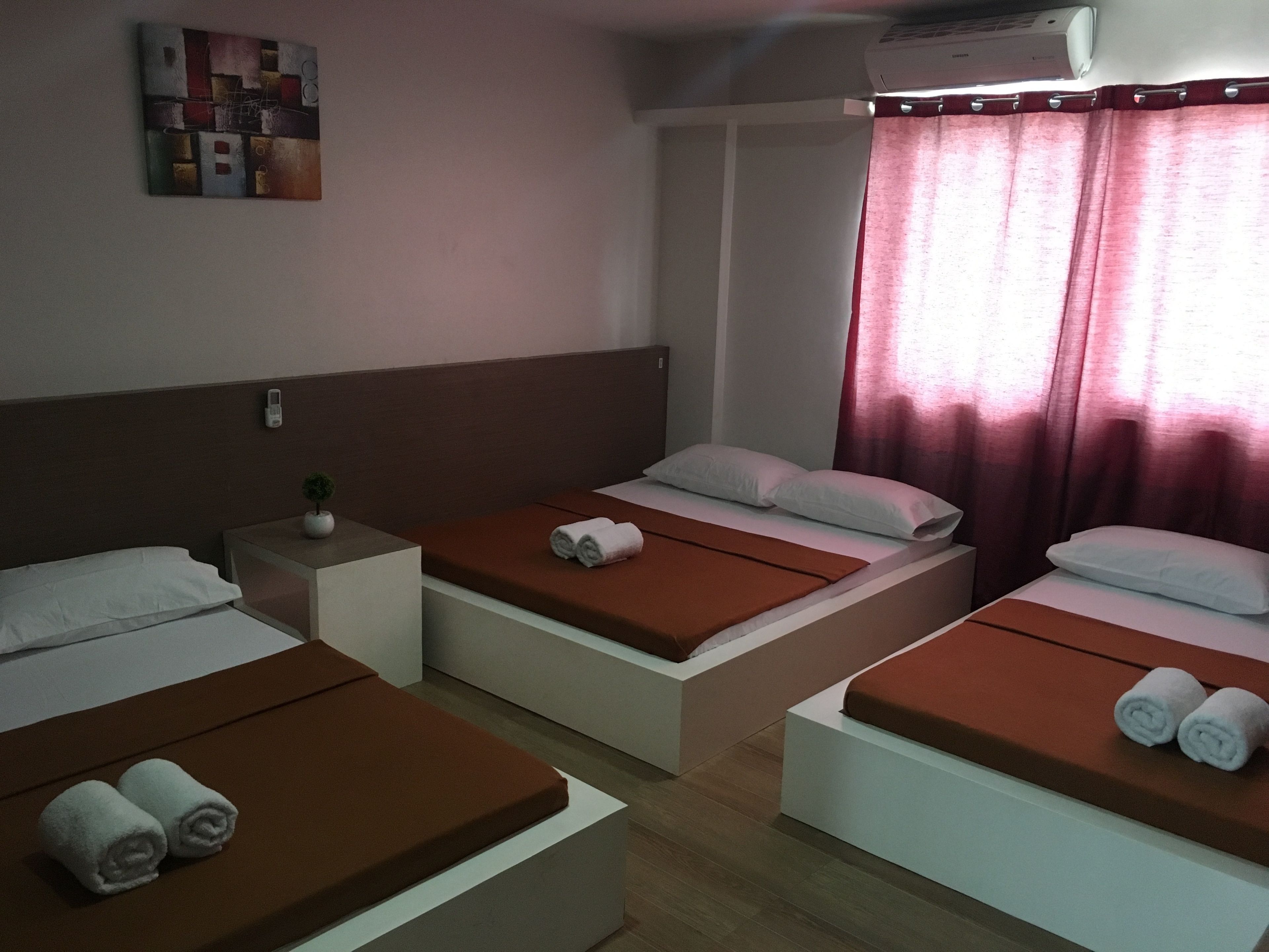 Une chambre d’hôtel avec trois lits simples, chacun doté d’un matelas marron et d’oreillers blancs, ainsi qu’une petite table de chevet agrémentée d’une plante.