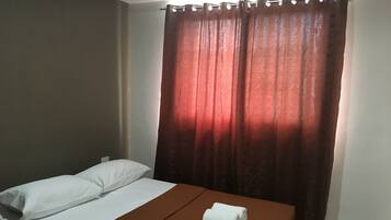 A single bed with white pillows and a brown blanket, a white towel on the bed, and a red curtain.