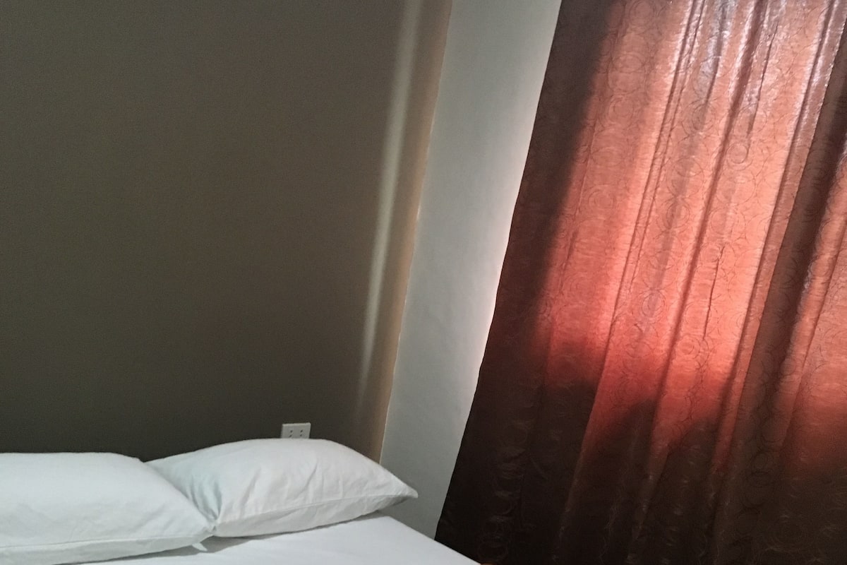 A single bed with a brown bedspread and white pillows, a folded white towel on the bed, and a window with red curtains.