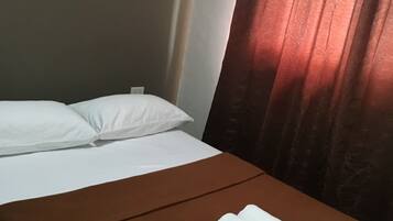 A single bed with a brown bedspread and white pillows, a folded white towel on the bed, and a window with red curtains.