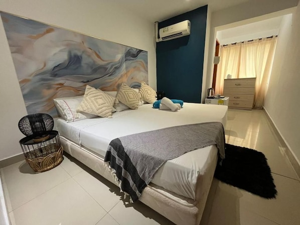 A bedroom with a bed, a wall-mounted air conditioner, a bedside table, a lamp, and a painting.