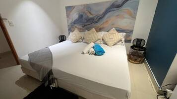 Een slaapkamer met een bed, kussens, een deken, een zwart vloerkleed, een kunstwerk aan de muur en een stoel.