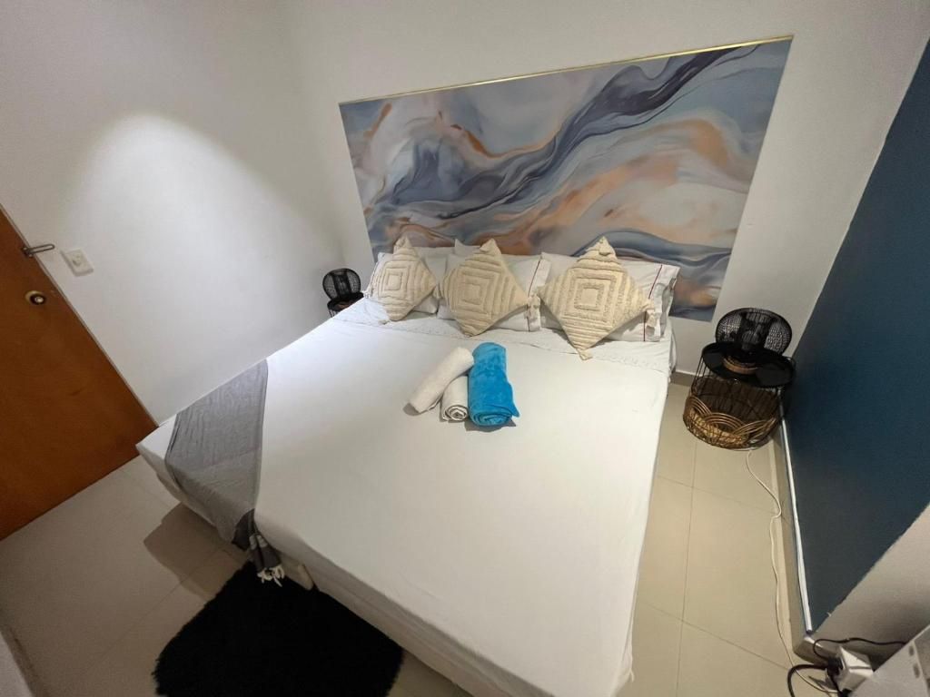 Ein Bett mit weißer Matratze, einem blauen Handtuch und Zierkissen. Darüber an der Wand hängt ein Gemälde.