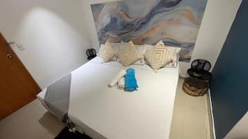 A bed with a white mattress, a blue towel, and decorative pillows. There is a painting on the wall above the bed.