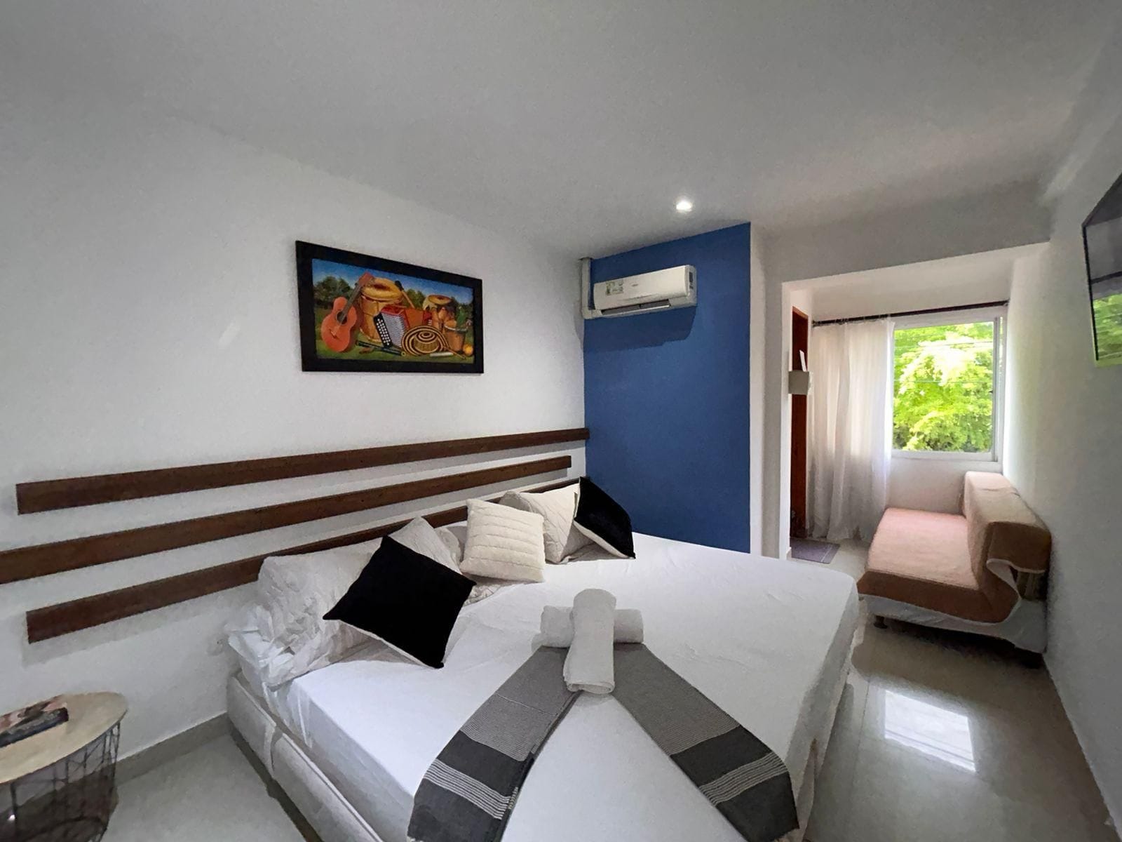 A hotel room with a bed, a painting, an air conditioning unit, a window with curtains, and a small table.