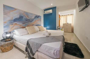 A bedroom with a bed, a large abstract painting, a standing lamp, a wicker basket, and a dresser. - Sweet Caribbean Hotel (San Andrés)