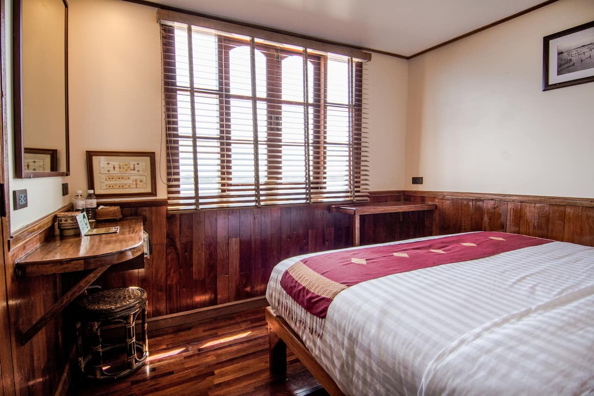 A room with a wooden floor, a bed with a white and maroon bedspread, a wooden desk with a chair, and a window with wooden blinds.