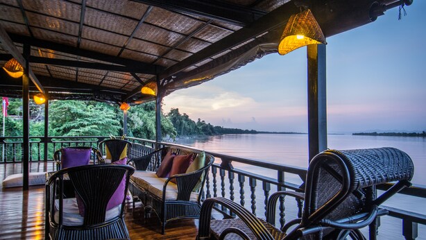 A covered outdoor seating area with wicker chairs and tables, overlooking a calm body of water and greenery. - The Vat Phou Cruise (Pakse)