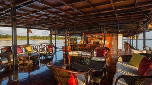 A covered deck with wicker furniture, a bar area, and a view of the water. - The Vat Phou Cruise (Pakse)