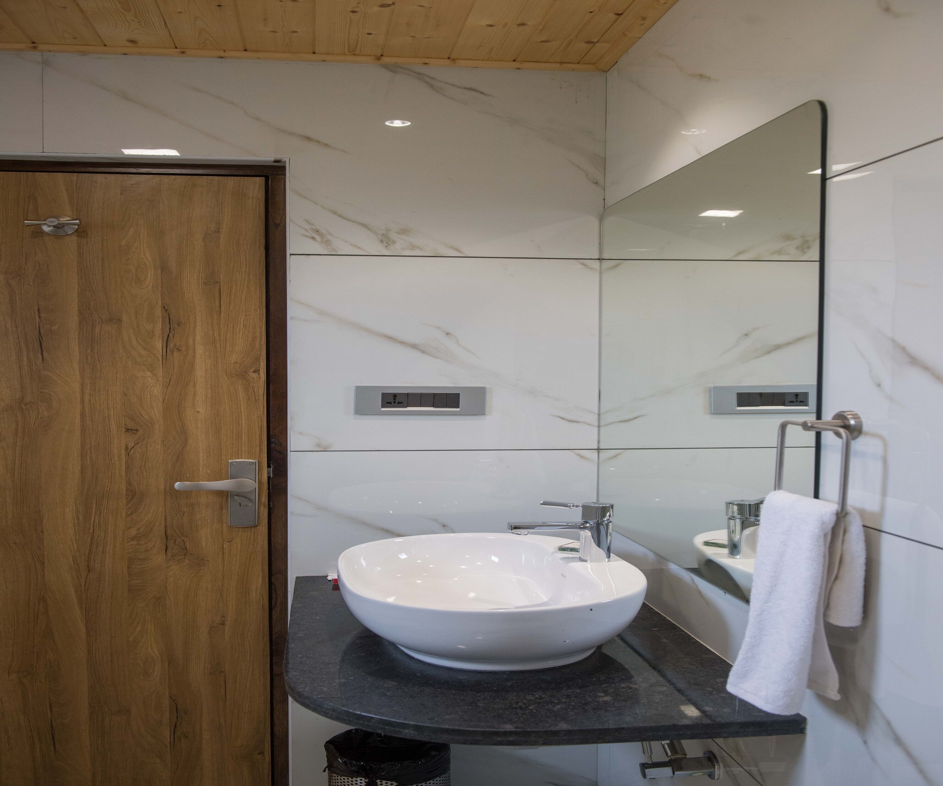 A modern bathroom with a wooden door, a white marble wall, a large mirror, a white sink, and a towel rack.