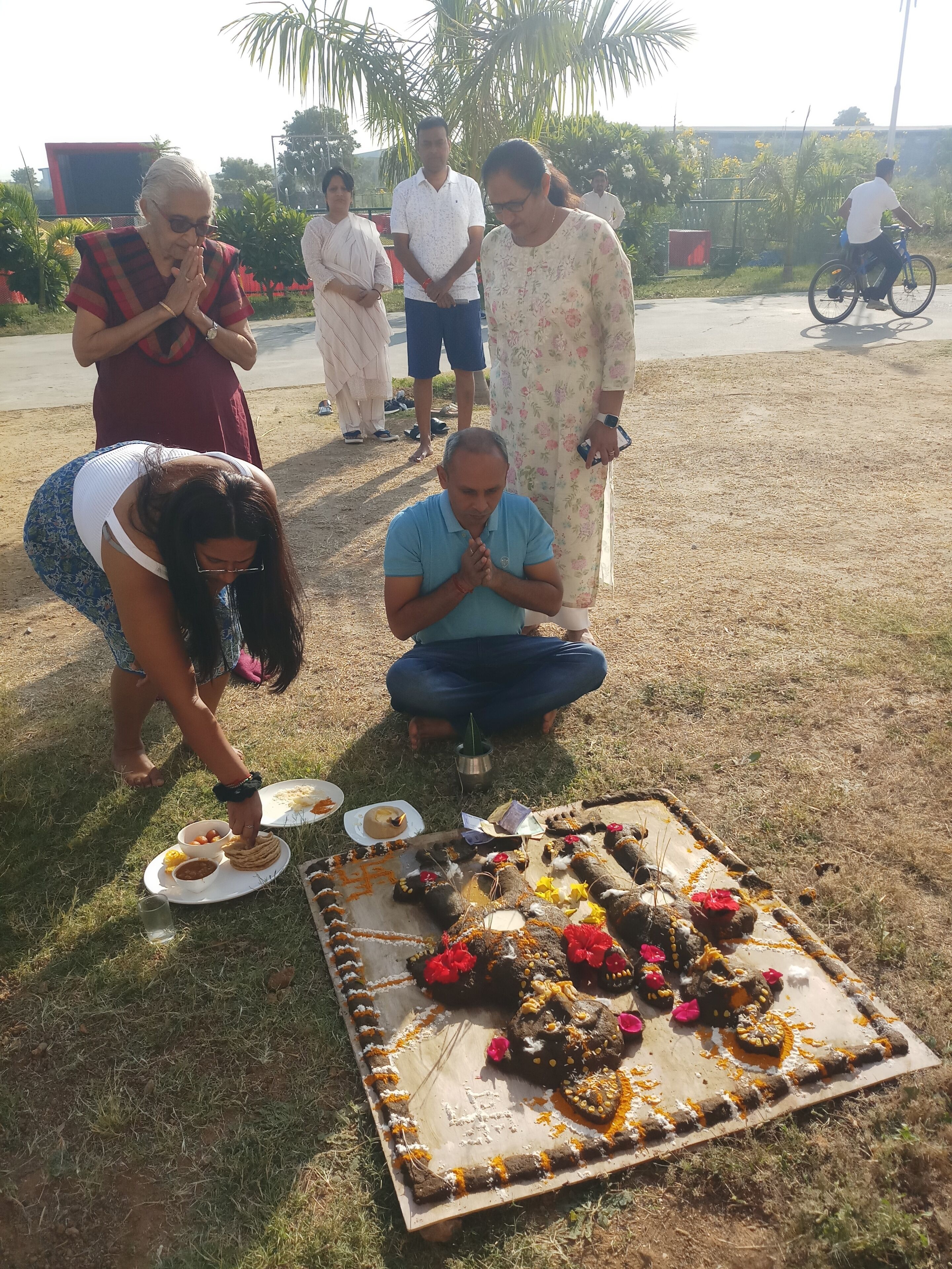 Un grupo de personas reunidas alrededor de un tapete decorado con comida y flores, al aire libre.