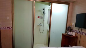 Quarto de hotel com porta deslizante para área de chuveiro, uma cama, uma pequena mesa com um controle remoto e uma televisão na parede.