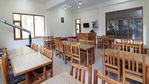 A dining area with multiple wooden tables and chairs, ceiling fans, and large windows. - Hotel Tashila - Manali (Manali)
