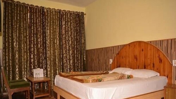 A wooden-framed bed with a white mattress and a headboard, a wooden nightstand, a chair with a green cushion, and a curtain with a leaf pattern. - Hotel Tashila - Manali (Manali)