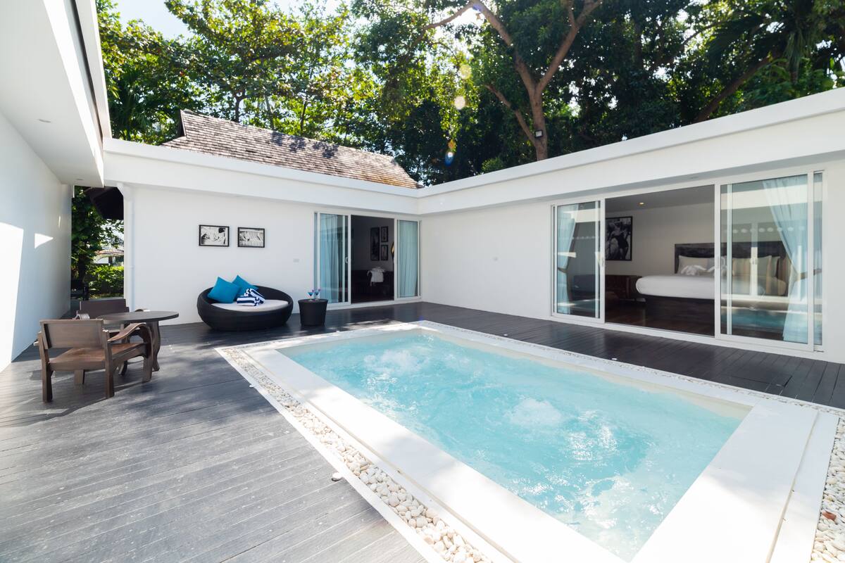 A modern outdoor pool area with a wooden deck, seating, and a glass-enclosed bedroom.