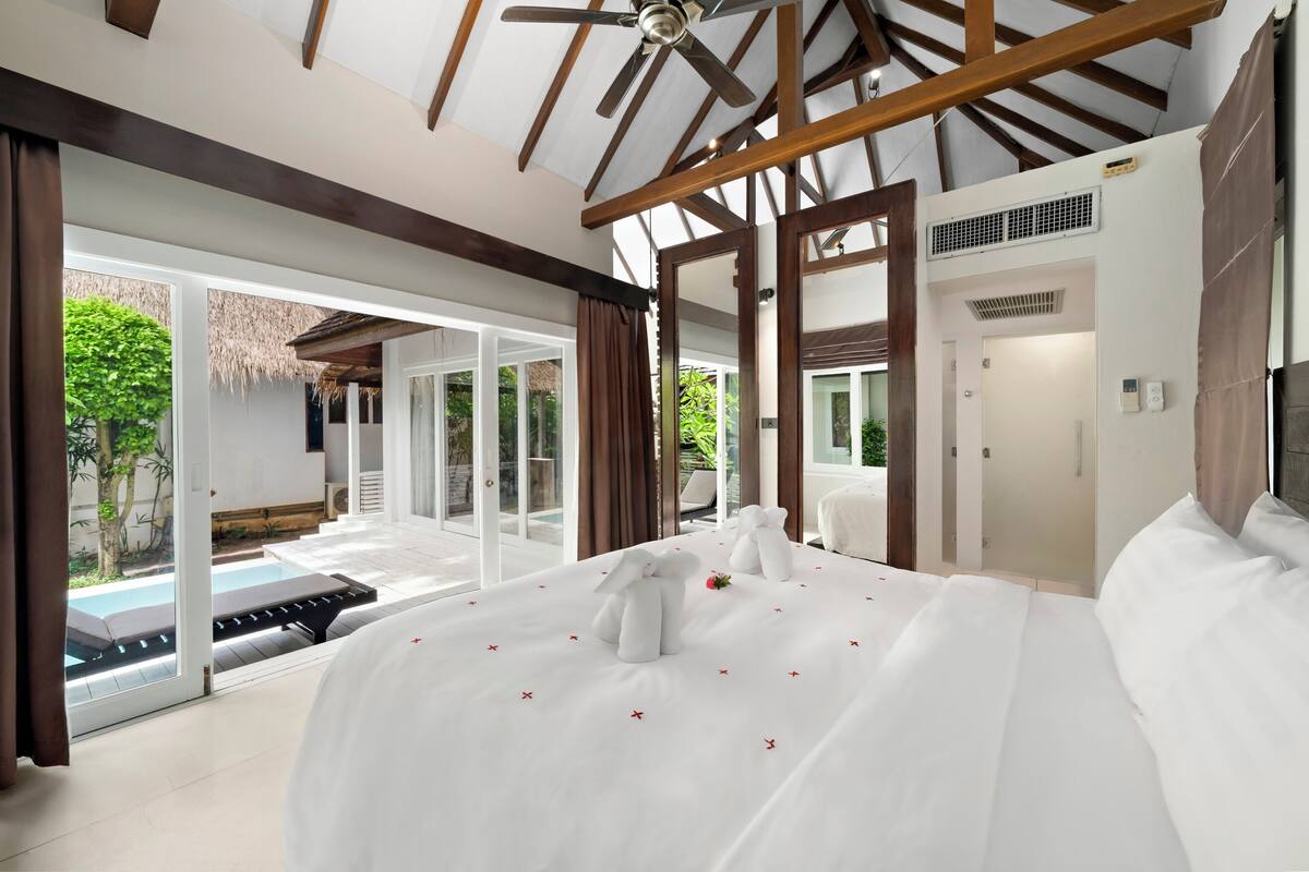 A spacious bedroom with a large bed, a ceiling fan, and a view of a swimming pool and garden.