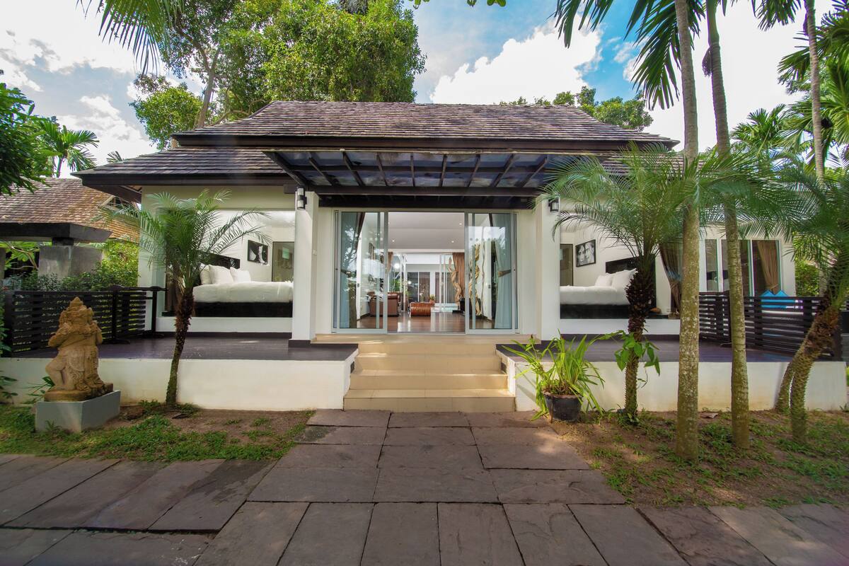 A modern house with a covered entrance, a stone statue, and a garden with palm trees.