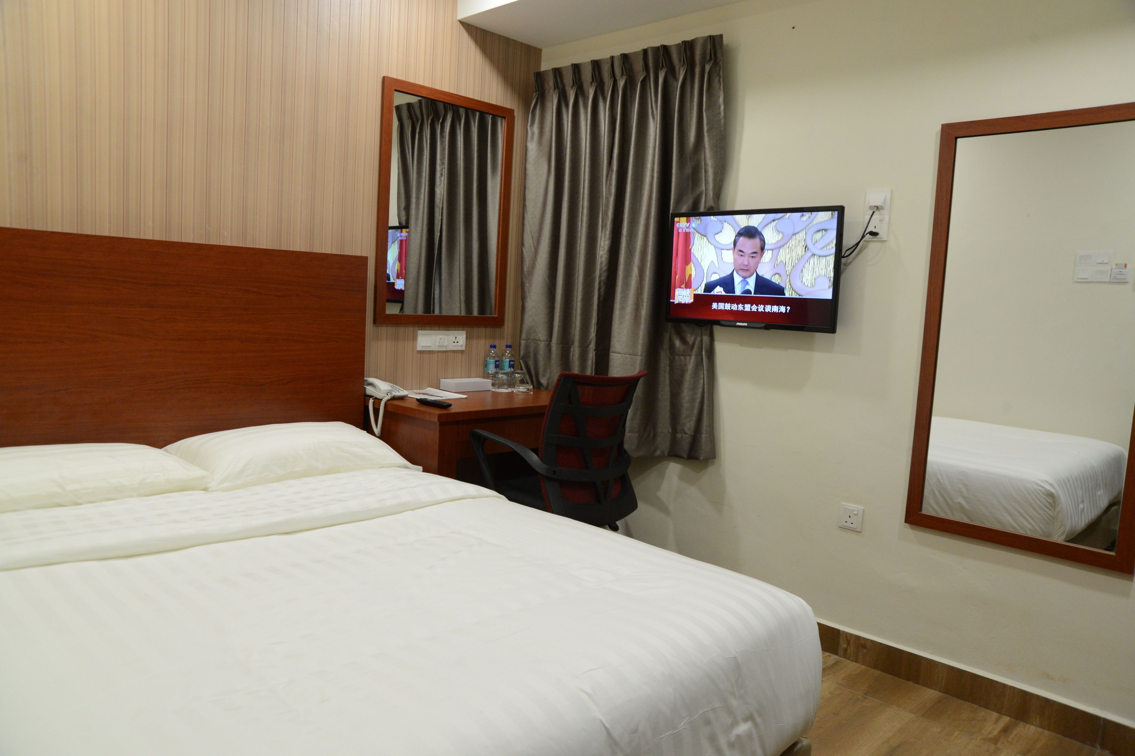 A hotel room with a bed, a desk with a chair, a television mounted on the wall, and a mirror.