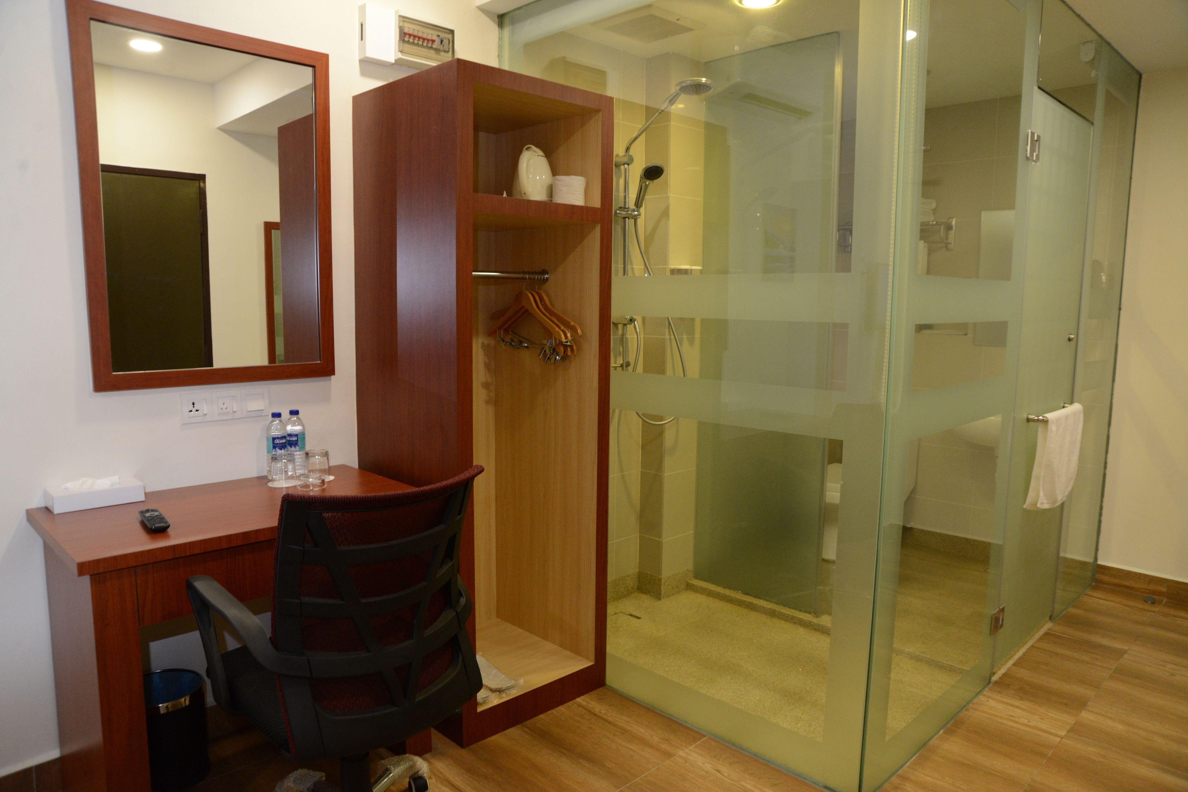 A hotel room with a wooden desk, a chair, a mirror, a wardrobe, and a glass-enclosed shower area.