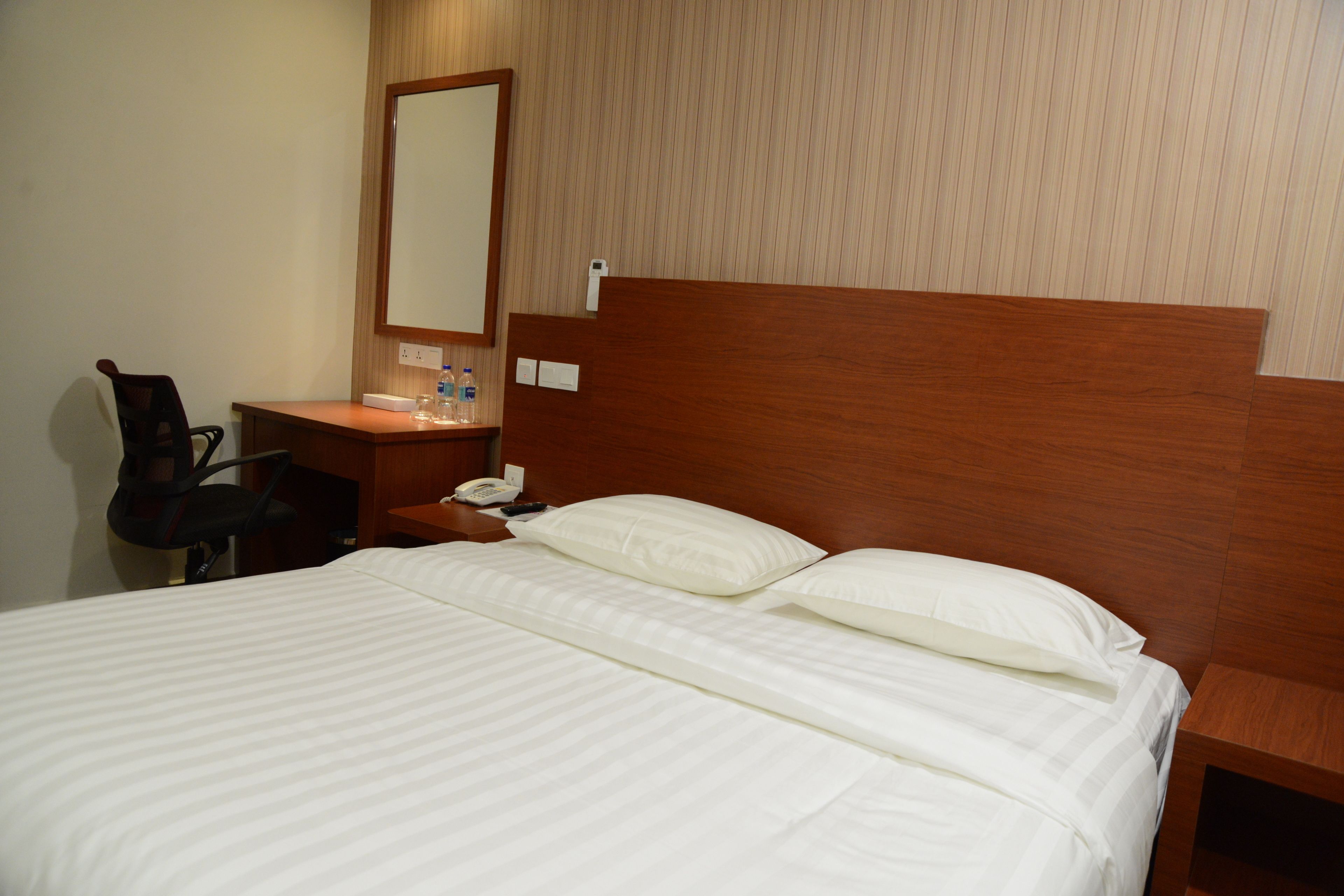 Habitación de hotel con cabecera de madera, cama con ropa blanca, mesita de noche con teléfono y botellas de agua, y espejo en la pared.