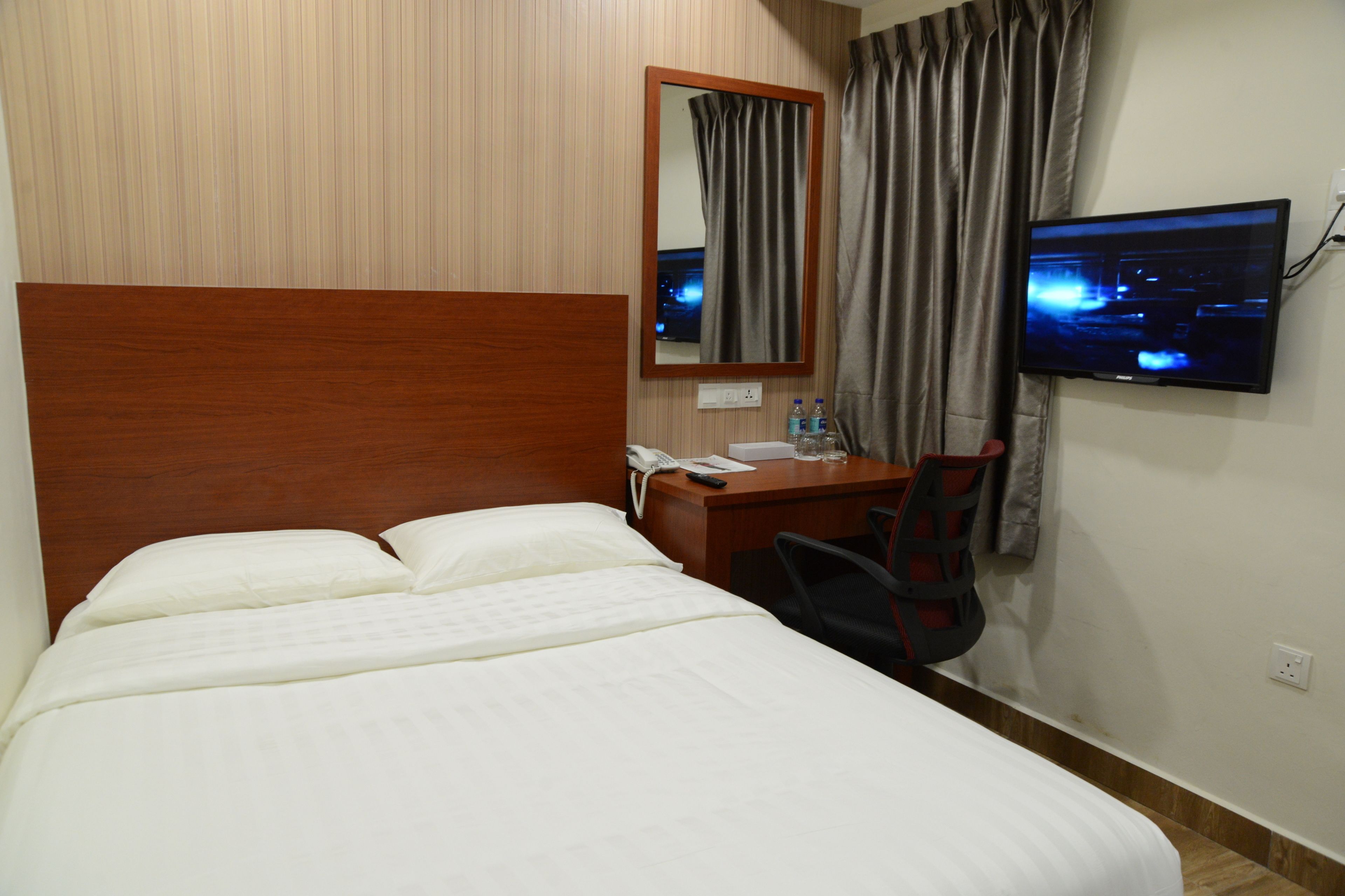 A hotel room with a bed, a desk, a chair, a TV, a mirror, and a window with curtains.