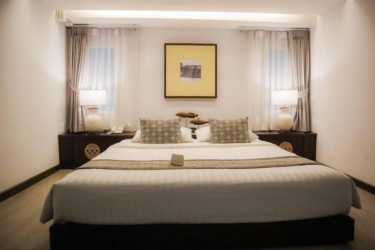 Habitación de hotel ordenada con una cama grande, dos mesitas de noche con lámparas, un cuadro enmarcado en la pared y cortinas en las ventanas.