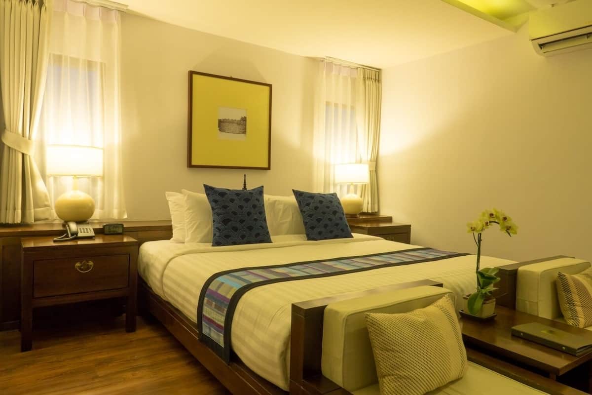Habitación de hotel con una cama grande, dos mesitas de noche con lámparas, un teléfono en la mesita y un cuadro en la pared, además de una pequeña mesa con una planta.