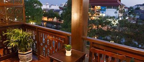 A wooden balcony with a table and chairs, a potted plant, and a view of buildings and trees.