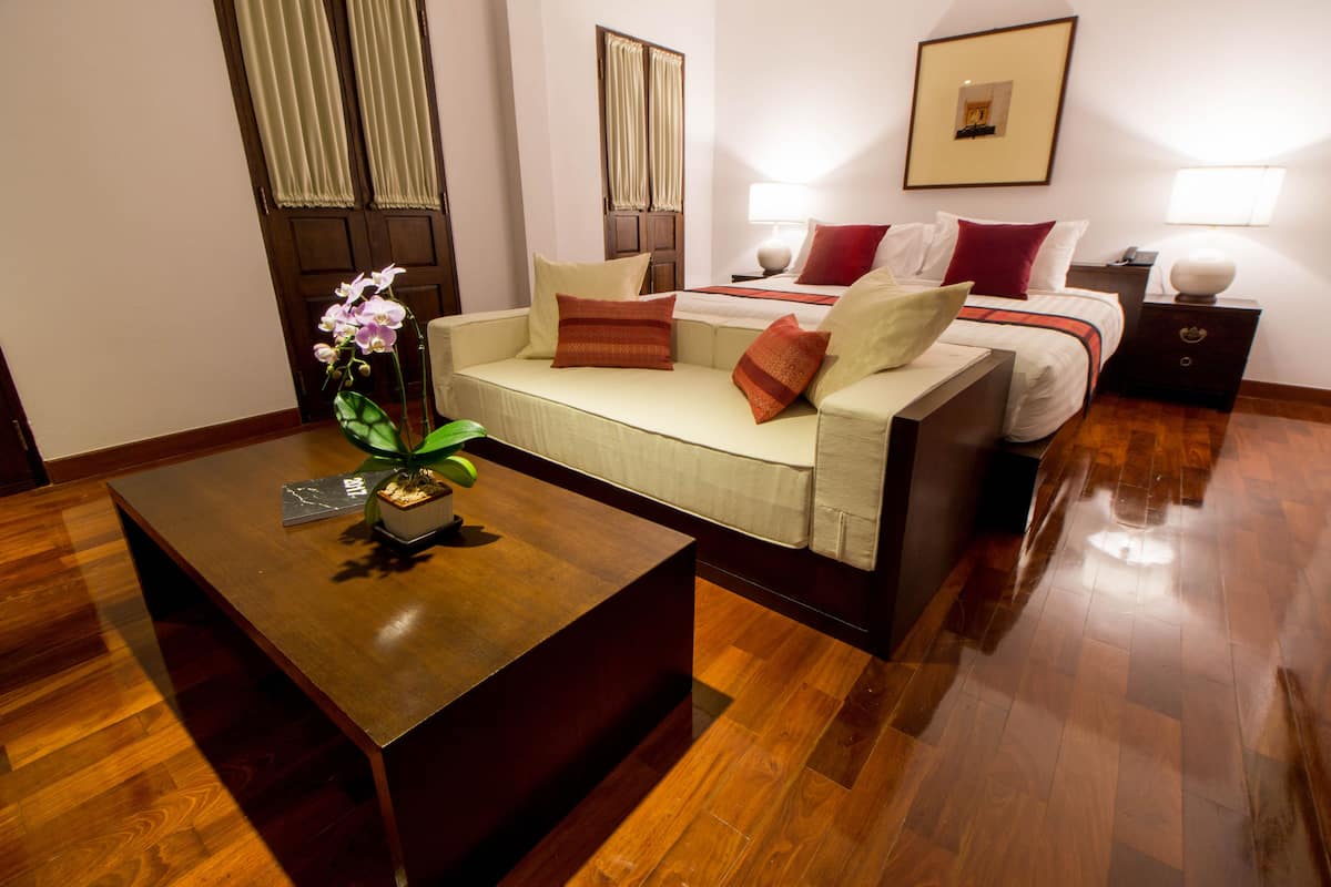 Habitación de hotel con suelo de madera, sofá, cama, mesita de noche, lámpara y un cuadro enmarcado en la pared.