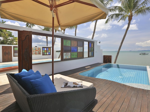 Un área junto a la piscina con terraza de madera, sofá de mimbre con cojines azules, una mesa con un libro, y vista al mar y palmeras.