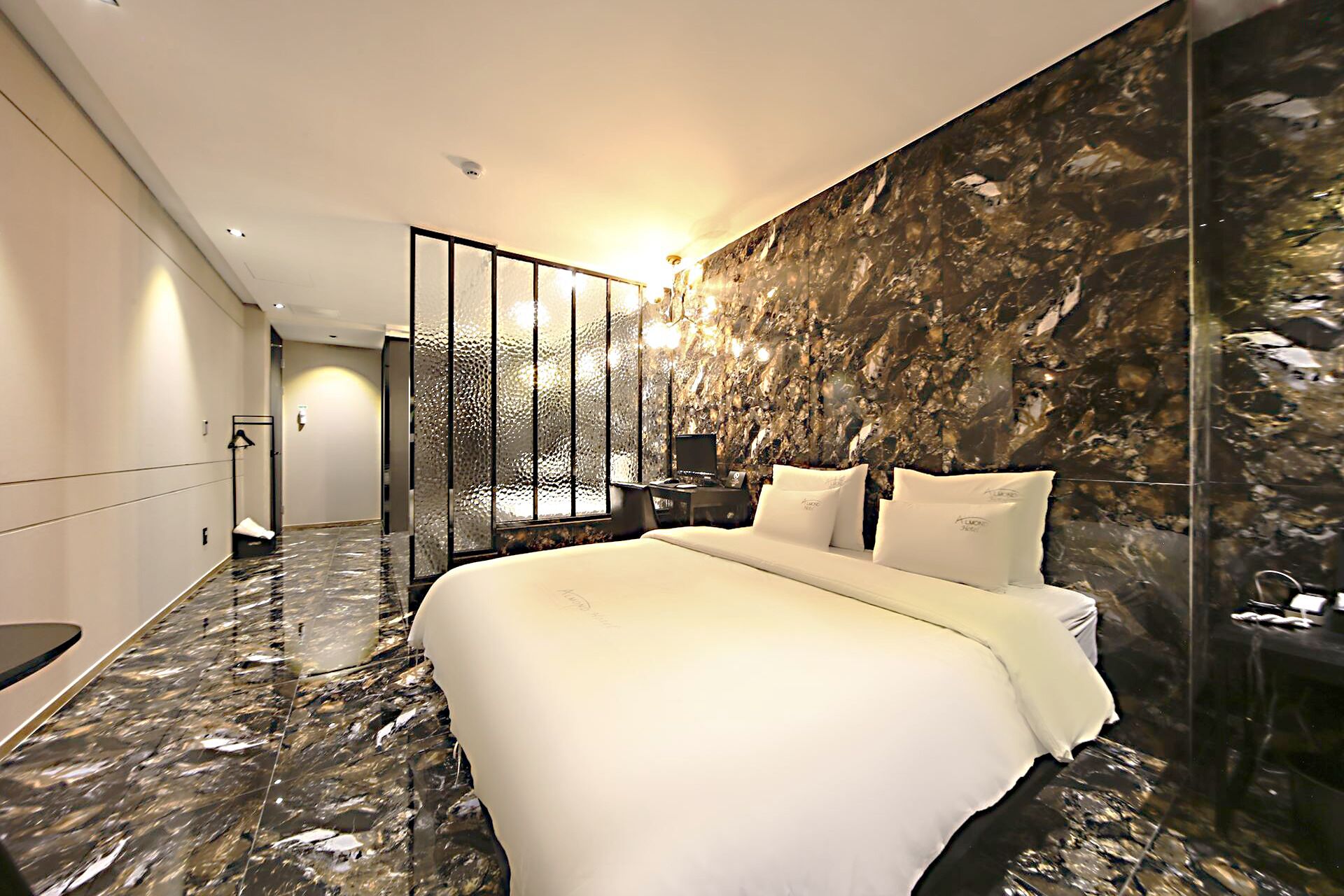 A modern hotel room with a large bed, a marble floor, a decorative wall, and a nightstand with a lamp.