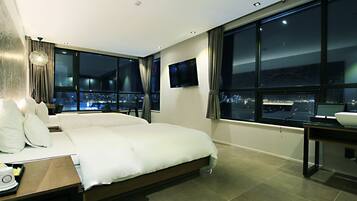 A modern hotel room with a large bed, a desk with a chair, a flat-screen TV, and a view of the city at night.