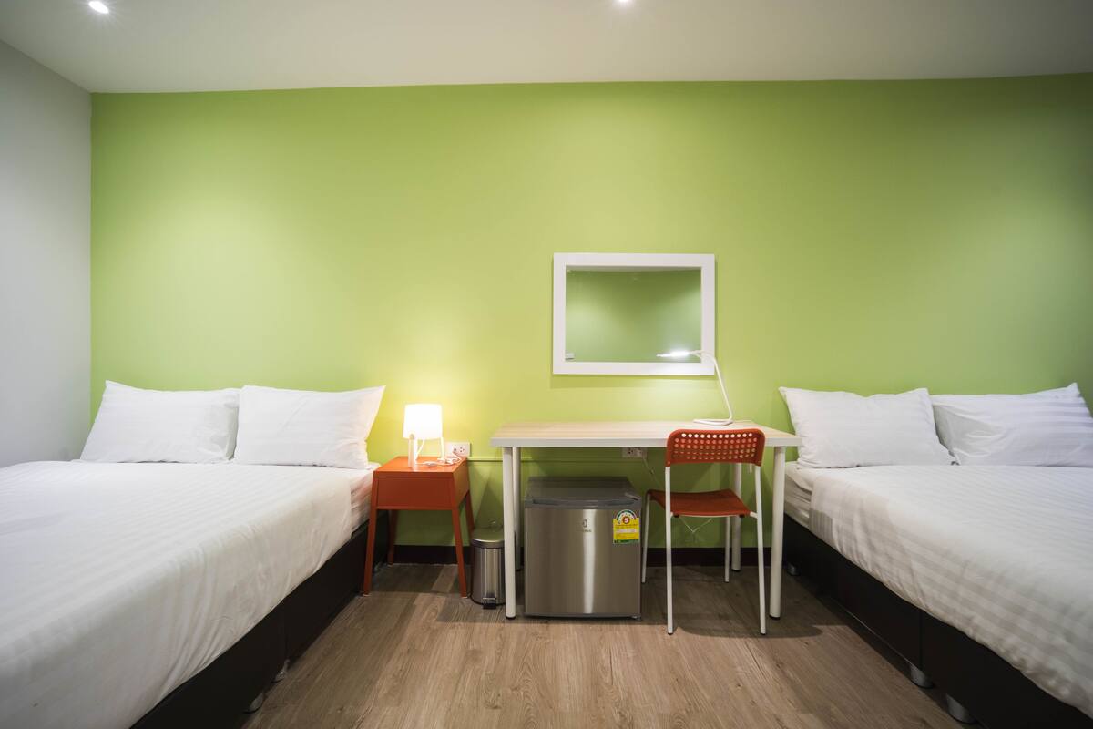A hotel room with two beds, a desk, a chair, a trash bin, and a mirror on the wall.