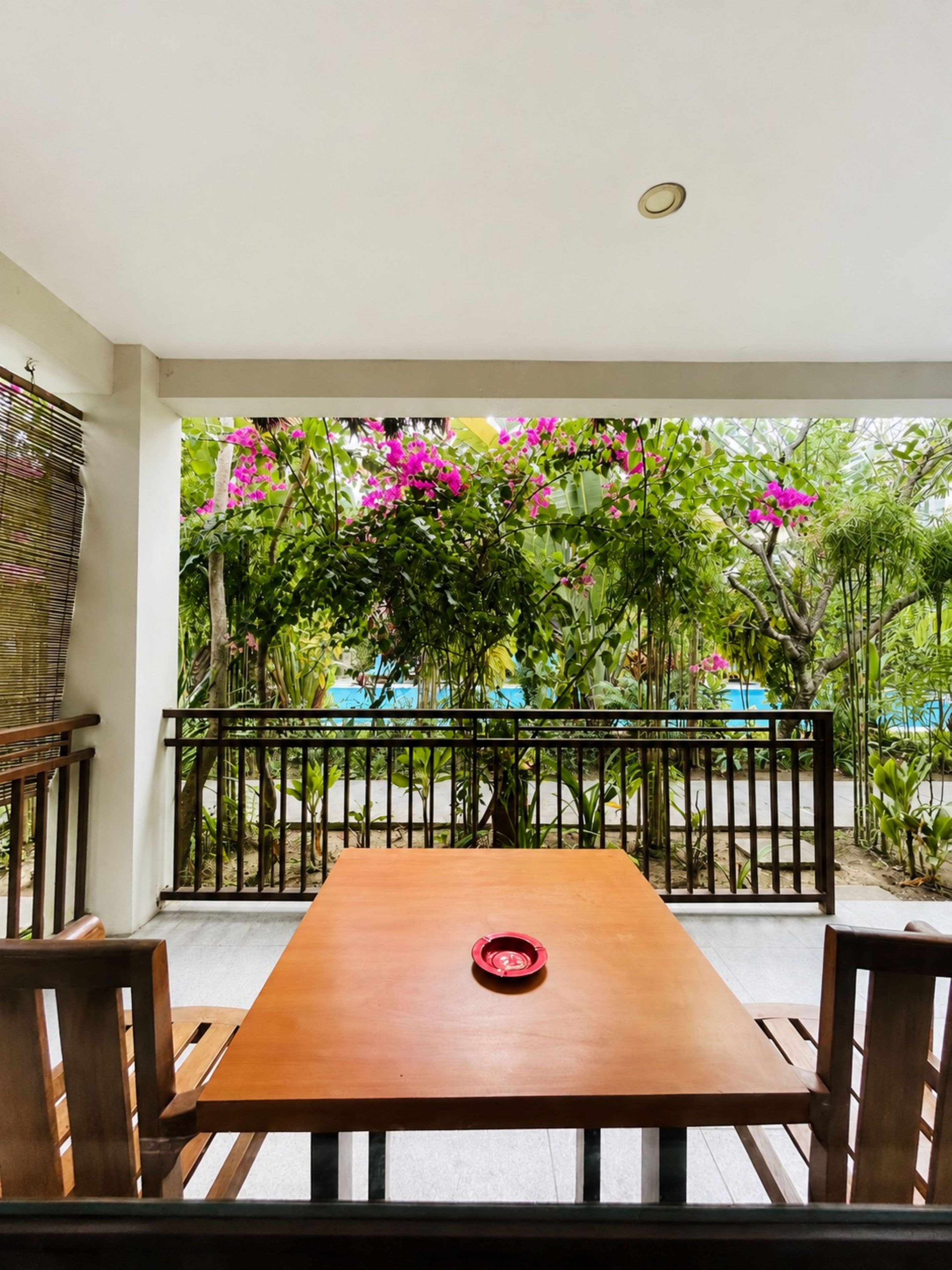 A wooden table with four chairs, a red object on the table, and a view of a garden with pink flowers and a black railing.