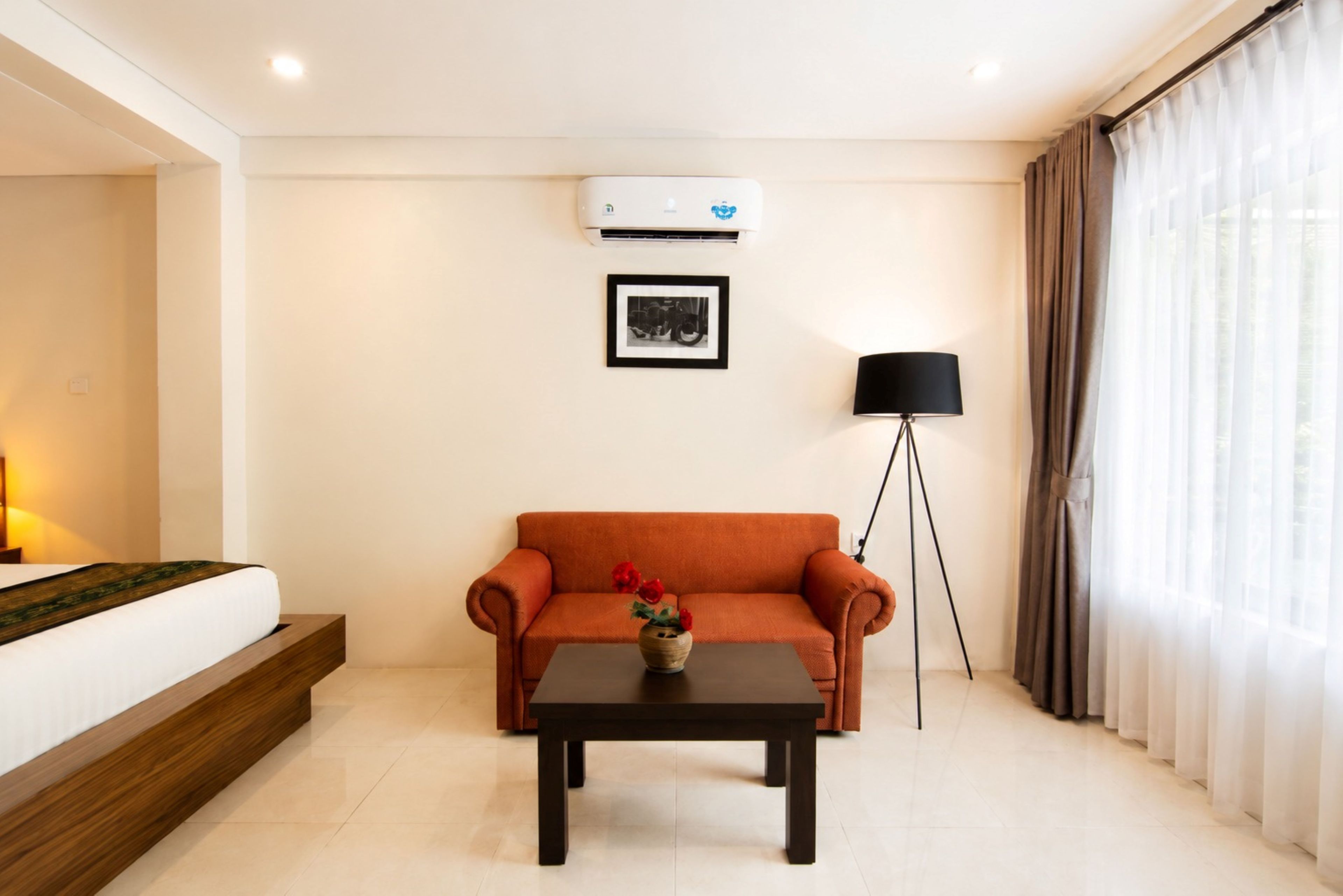 A hotel room with a bed, a sofa, a small table, a floor lamp, and an air conditioning unit.