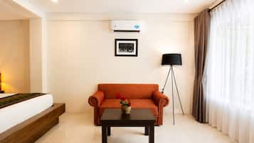 A hotel room with a bed, a sofa, a small table, a floor lamp, and an air conditioning unit.