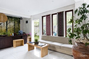 A modern living room with a sofa, a coffee table, and a desk with a computer. There are large windows with curtains, a hanging light fixture, and a potted plant.