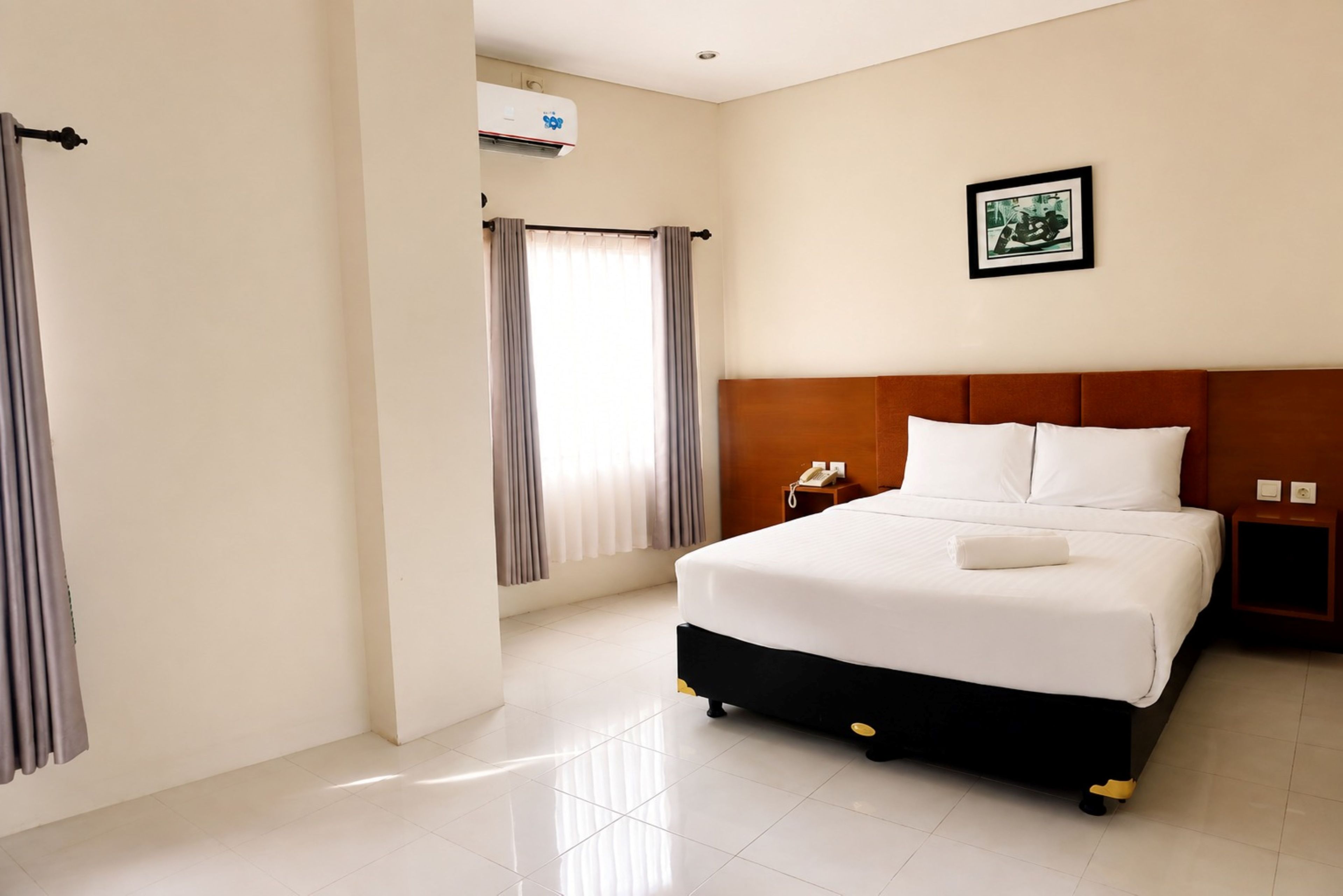 A hotel room with a bed, a telephone, a picture on the wall, and an air conditioning unit.