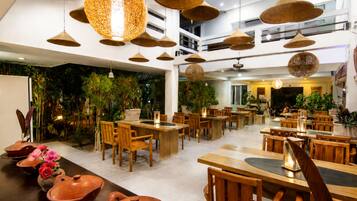 A restaurant with multiple dining tables and chairs, hanging pendant lights, and indoor plants.